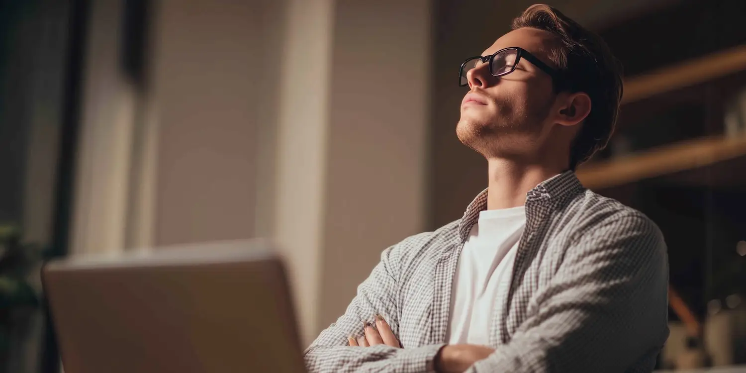 young man wearing glasses leaning back with arms folded taking a mindful break from his laptop